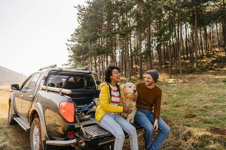 A couple are sitting on their SUV in nature with their dog.